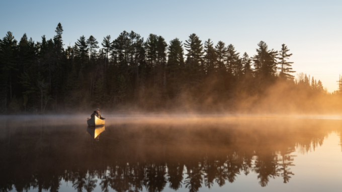 a fly angler wearing a cap and fishing jacket casts a fly line from a canoe on a lake